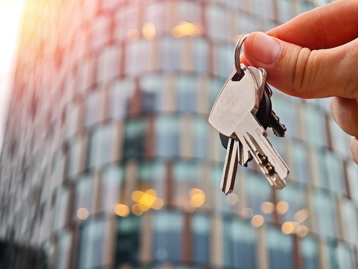 Close-up of hand holding a house key and keychain in front of an apartment building, concept of homeownership