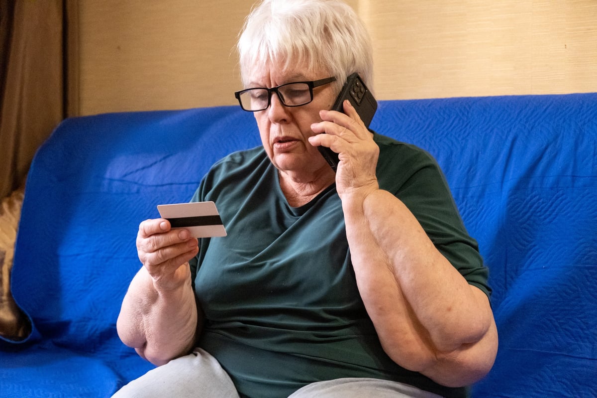 Senior woman in glasses sitting on sofa looking on credit card and talking on phone. Concept of scam, money loss, banking protection, finance fraud, customer service, remote payment