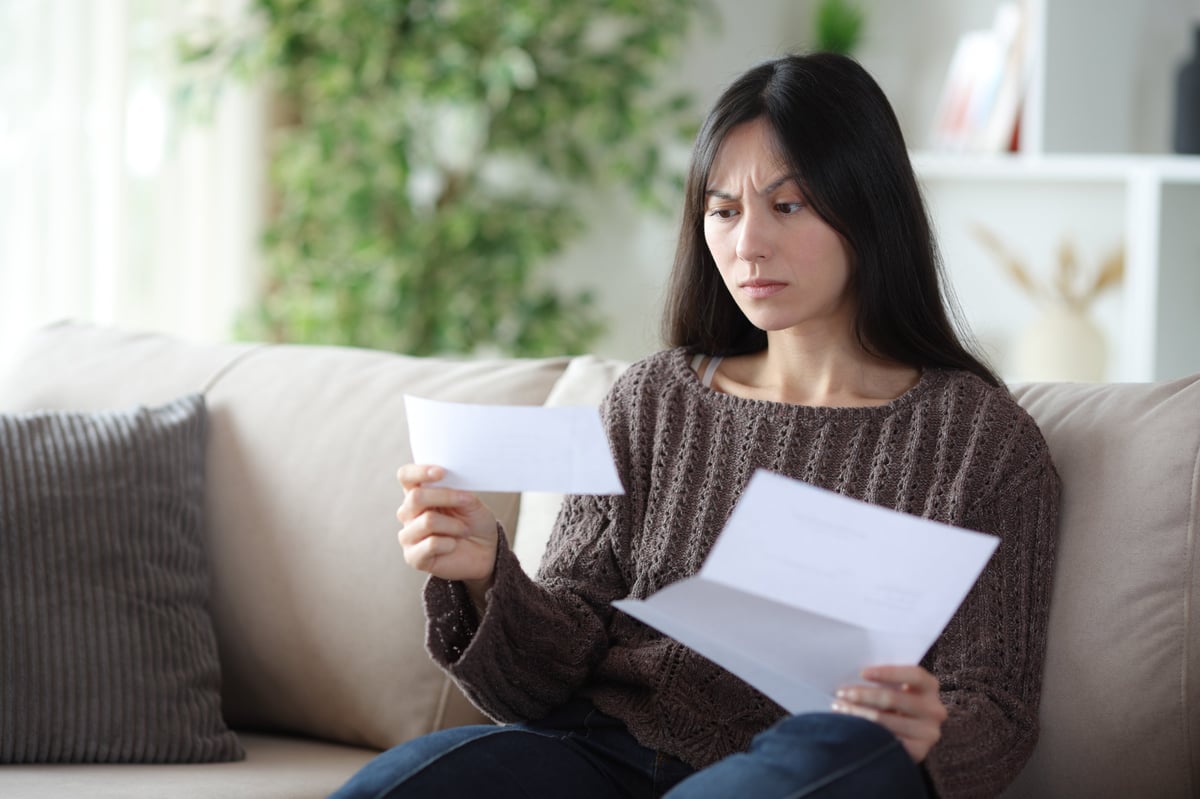 Worried asian woman reading bad news in a letter sitting on a couch at home