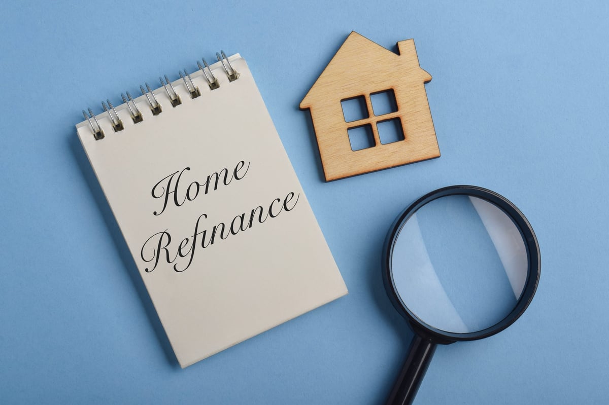 Notepad with home refinance text, house icon and magnifying glass on blue background. Concept of mortgage refinancing, home loan review, property finance, lending and housing investment.